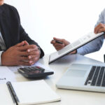 A man discussing some numbers with another on a paper and laptop
